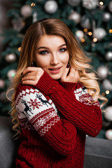 Charming long-haired blonde in a sweater with deers poses during Christmas time near the Christmas tree. New Year photo session of a sexy blonde in a sweater