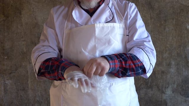 elderly man with gray beard in kitchen suit and white apron wears thin gloves before working with meat