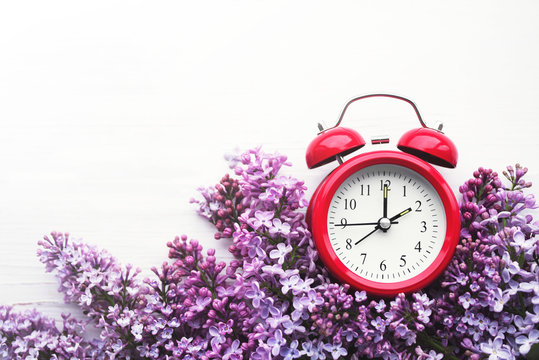 Red alarm clock and bouquet of pink lilacs on a white surface, top view. Hello spring, spring time, conceptual image