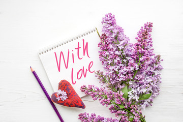 Text WITH LOVE on a white open notebook, red heart and bouquet of lilacs on a white wooden surface,...