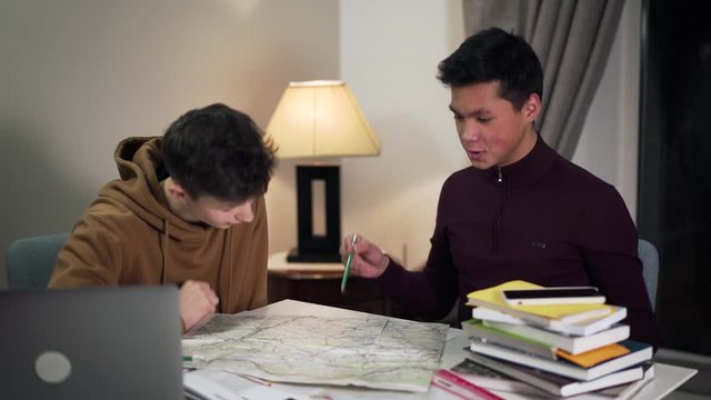 Camera approaching to excited college students discussing geography and giving dap. Happy Asian and Caucasian boys sitting at the table and talking. Lifestyle, education, enjoyment.