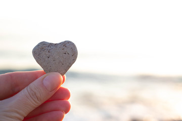 female hand holds a heart of natural stone against the background of the sea in the sun. Reminder...