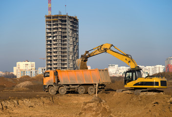 Excavator load the sand to the dump truck on construction site. Backhoe digs the ground for the foundation and construction of a new building. Background of the tower cranes