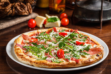 Obraz na płótnie Canvas Italian food. Classic thin pizza with large sides, prosciutto, cherry tomatoes, arugula, parmesan cheese. Serving dishes in a restaurant on a white plate with red wine.