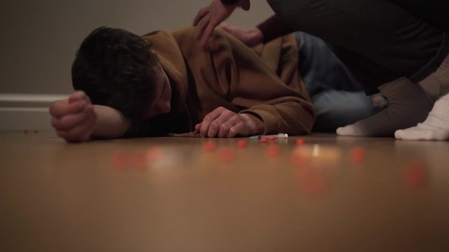 Portrait of addicted junkie lying on the floor with pills and syringe scattered around. Unrecognizable man trying to wake narcomaniac up. Overdose, dependence, drug addiction.
