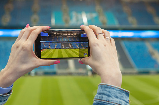 A girl in a football (soccer) stadium with a smartphone takes a picture of the green field of the arena. Modern technology at sports events.