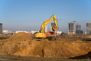 Excavator load the sand to the dump truck on construction site. Backhoe digs the ground for the foundation and construction of a new building. Background of the tower cranes