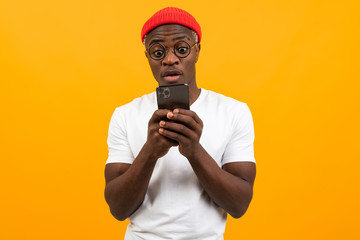 handsome african man in a white t-shirt with a red hat communicates on a social network in a smartphone on a yellow studio background