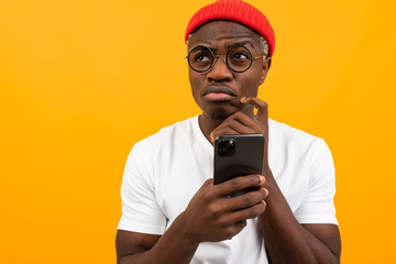 Attractive pensive charming black man in a white T-shirt with a smartphone in his hands on a yellow studio background with copy space