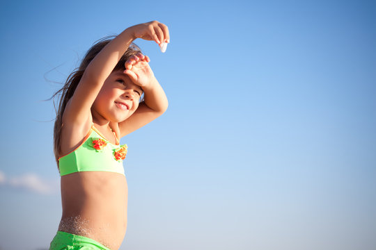 Small smiling girl in bright swimwear standing in sunshine and looking at little stone with blue sky background on clear sunny summer day. Travelling, vacations, relaxation, family weekend concept