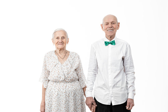 Portrait of happy, smiling, romantic senior man with his wife, old woman in white dress and old man in white shirt holding hands together and looking at the camera, isolated over white background