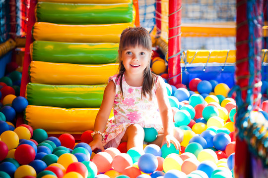 Small smiling girl in dress sitting in colorful soft decorative balls in playroom with sunshine from aside. Happy childhood and active children concept