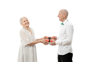 Old happy smiling woman in white dress gives a gift to her husband in white shirt and looking at the camera isolated over white background, new year, happy birthday, St. valentine's day celebration