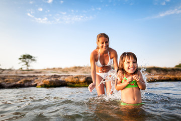 Fototapeta premium Young blond woman in white bikini and small happy girl having fun in sea water with rocky beach at background on clear summer day. Travelling, vacations, relaxation, family weekend concept