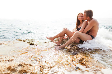 Young happy beautiful couple woman and man sitting on rocks and looking at each other with sea at background on clear summer day. Travelling, vacations, romantic weekend, honeymoon concept