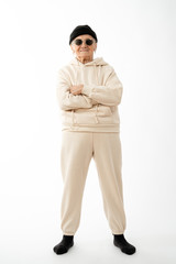 Fototapeta premium Stylish old hipster woman in beige hoodie black hat and sunglasses stands with crossed hands and looking at the camera isolated over white background