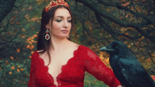 Portrait pretty brunette woman with huge black raven on hand. Image of queen in red dress, elegant hairstyle with gold crown and ruby earrings. Posing for camera. Background trees with yellow leaves