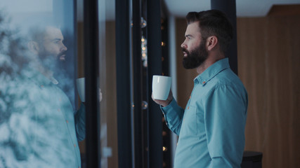Close up young handsome man bearded drinking coffee and enjoying view from window relax handsome male tea window apartment attractive caucasian human face relaxation adult panorama indoors slow motion