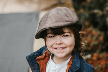 Closeup portrait of cute little smiling toddler boy with long hair in brown cap outdoors. Retro or vintage style.  Copy space.