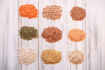 piles of various legumes (chickpeas, green peas, red lentils, buckwheat, quinoa, chia seeds, bulgur, yellow peas, green mung beans) onwhite wooden background