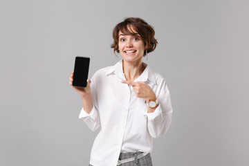 Fototapeta premium Pretty business woman in white shirt posing isolated on grey background. Achievement career wealth business concept. Mock up copy space. Pointing index finger on mobile phone with blank empty screen.