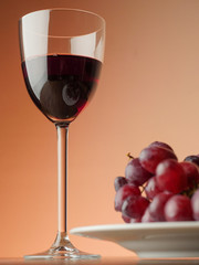 Glass of red wine and plate with red grapes on a wooden table, Warm light background. Glass is in focus. Low angle of view.