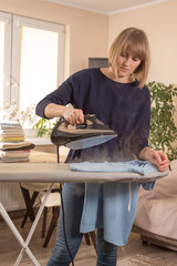 Kobieta trzyma żelazko z którego wydostaje się para wodna. Prasowanie odzieży na desce do prasowania. Wyprasowane ubrania leżą na stole w tle.