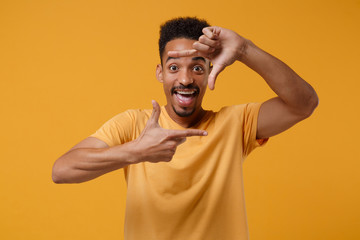 Fototapeta na wymiar Excited funny young african american guy in casual t-shirt posing isolated on yellow orange wall background in studio. People lifestyle concept. Mock up copy space. Making hands photo frame gesture.