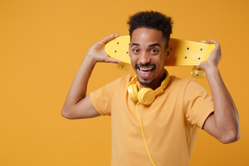 Excited young african american guy in casual t-shirt, headphones posing isolated on yellow orange wall background studio portrait. People lifestyle concept. Mock up copy space. Holding skateboard.