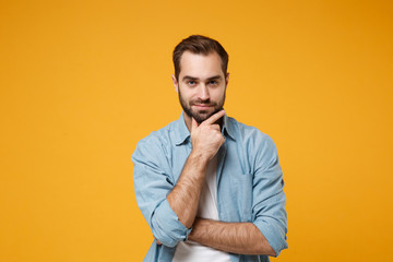 Attractive young bearded man in casual blue shirt posing isolated on yellow orange background studio portrait. People sincere emotions lifestyle concept. Mock up copy space. Put hand prop up on chin.