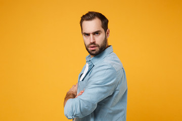 Obraz premium Side view of serious young bearded man in casual blue shirt posing isolated on yellow orange background in studio. People lifestyle concept. Mock up copy space. Holding hands crossed, looking camera.