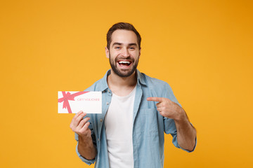 Laughing young man in casual blue shirt posing isolated on yellow orange wall background, studio portrait. People lifestyle concept. Mock up copy space. Pointing index finger on gift certificate. - obrazy, fototapety, plakaty