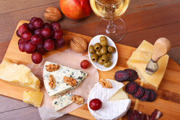 Assortment of cheese with fruits, grapes, nuts, glass with wine and cheese knife on a wooden serving tray.