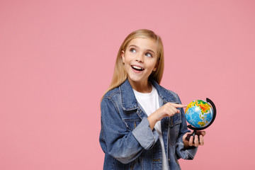 Pensive little blonde kid girl 12-13 years old in denim jacket posing isolated on pastel pink background. Childhood lifestyle concept. Mock up copy space. Pointing index finger on Earth world globe.