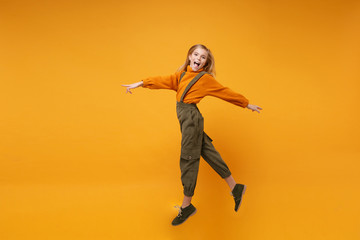 Excited little kid girl 12-13 years old in turtleneck, jumpsuit isolated on orange yellow wall background in studio. Childhood lifestyle concept. Mock up copy space. Jumping, spreading hands and legs.