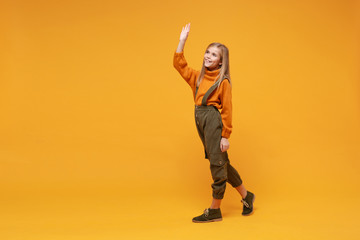Cute little blonde kid girl 12-13 years old in turtleneck jumpsuit isolated on orange yellow background. Childhood lifestyle concept. Mock up copy space. Waving greeting with hand as notices someone.