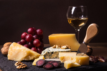 Assorted cheeses, nuts, grapes, fruits, smoked meat and a glass of wine on a serving table. Dark and Moody style. Free space for text.