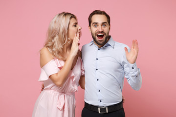 Excited young couple two guy girl in party outfit celebrating isolated on pastel pink background. People lifestyle Valentine's Day Women's Day birthday holiday concept. Whispering secret behind hand.