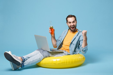Happy traveler tourist man in summer clothes isolated on blue background. Passenger travel on weekend. Air flight journey. Sit in inflatable ring work on laptop, hold beer bottle doing winner gesture.