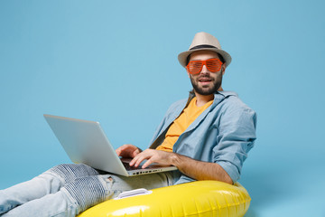 Traveler tourist man in yellow clothes eyeglasses isolated on blue background. Passenger traveling abroad on weekend. Air flight journey concept. Sit in inflatable ring work on laptop booking hotel.