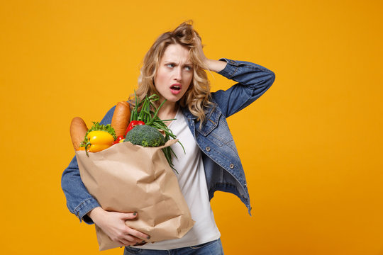 Exhausted woman in denim clothes isolated on orange background. Delivery service from shop or restaurant concept. Hold brown craft paper bag for takeaway mock up with food products put hand on head.