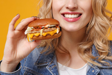 Cropped image of smiling young woman in denim clothes isolated on orange background studio portrait. Proper nutrition or American classic fast food concept. Mock up copy space. Holding in hand burger.