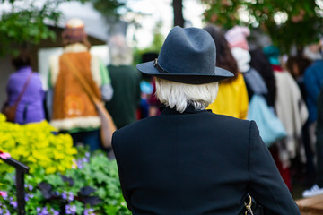 Rear view of woman with gray hair wearing hat and blue blazer while looking at defocus standing crowd in event during festival