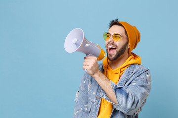 Cheerful young hipster guy in fashion jeans denim clothes posing isolated on pastel blue background studio portrait. People lifestyle concept. Mock up copy space. Looking aside, scream in megaphone. - obrazy, fototapety, plakaty