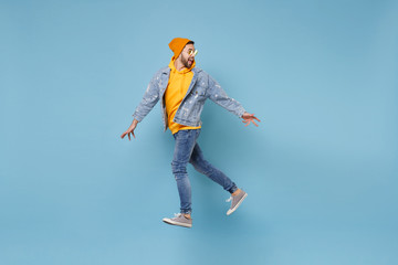 Side view of excited young hipster guy in fashion jeans denim clothes posing isolated on pastel blue background. People lifestyle concept. Mock up copy space. Jumping, spreading hands, looking aside.