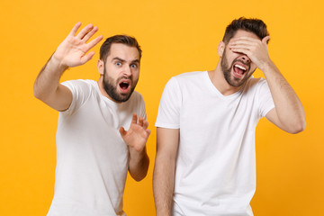 Shocked scared young men guys friends in white blank t-shirts posing isolated on yellow orange background. People lifestyle concept. Mock up copy space. Covering eyes with palm covering with hands.