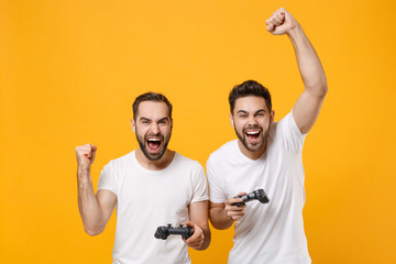 Cheerful young men guys friends in white t-shirts posing isolated on yellow orange background in studio. People lifestyle concept. Mock up copy space. Play game with joystick doing winner gesture. © ViDi Studio