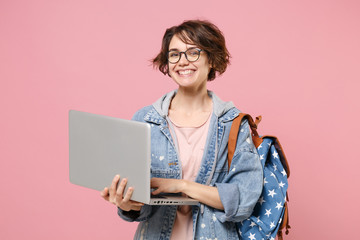 Cheerful young woman student in denim clothes glasses backpack isolated on pastel pink background. Education in high school university college concept. Mock up copy space. Work on laptop pc computer. - obrazy, fototapety, plakaty