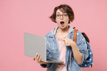 Shocked young woman student in denim clothes eyeglasses backpack isolated on pastel pink...