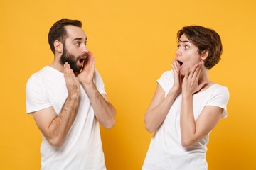 Amazed young couple friends bearded guy girl in white t-shirts posing isolated on yellow orange background. People lifestyle concept. Mock up copy space. Looking at each other, put hands on cheeks.